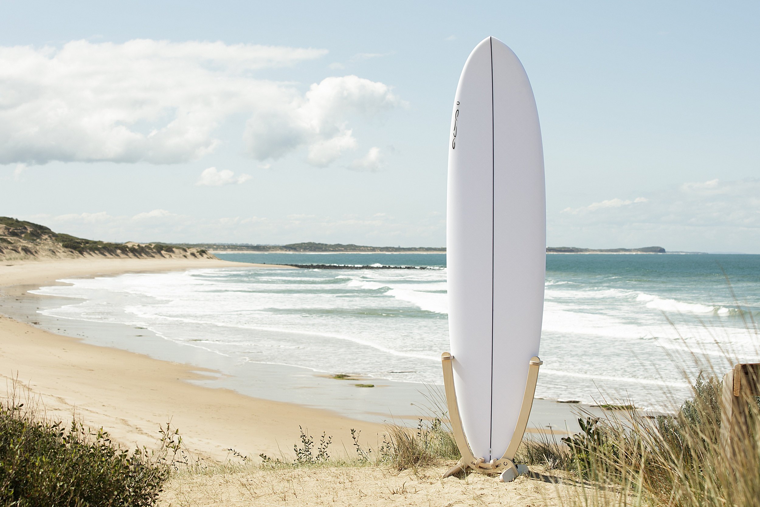An all white Surfboard with a GLYD O decal on its nose, is mounted in a GLYD O surfboard rack. The rack with board is standing on a sandy dune with the beach, waves and ocean in the back ground, coastal dunes in the distance. The Surfboard Rack is a Beautiful birchwood curved single surfboard rack in the shape of the tail of a surfboard, with hooks to hold the rails and tail and two slightly curved feet connected perpendicular to the upright curved arms. Image is taken from a slight angle in relations to the vertical surfboard rack. Rectangular image.