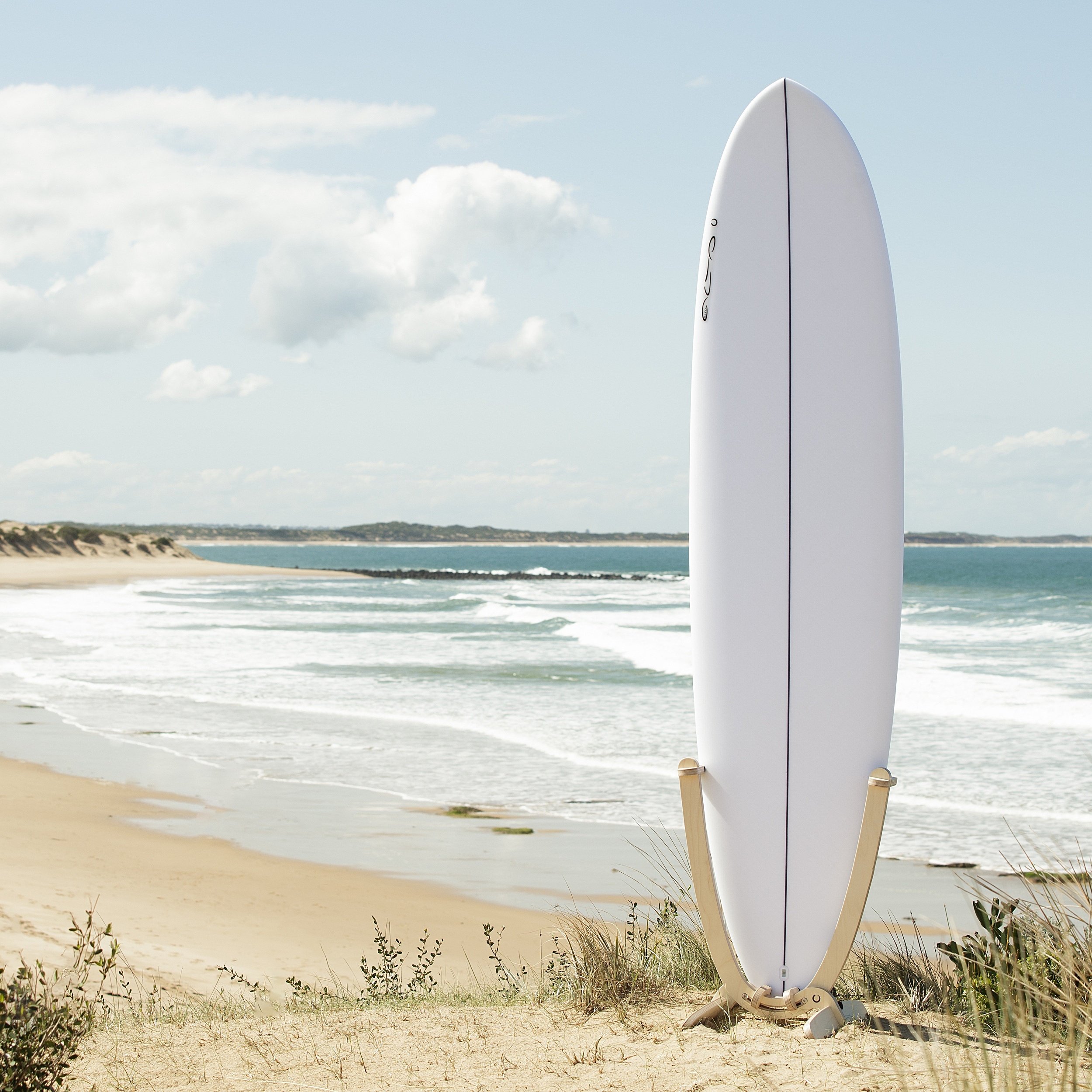 An all white Surfboard with a GLYD O decal on its nose, is mounted in a GLYD O surfboard rack. The rack with board is standing on a sandy dune with the beach, waves and ocean in the back ground, coastal dunes in the distance. The Surfboard Rack is a Beautiful birchwood curved single surfboard rack in the shape of the tail of a surfboard, with hooks to hold the rails and tail and two slightly curved feet connected perpendicular to the upright curved arms. Image is taken from a slight angle in relations to the vertical surfboard rack. Square Image.