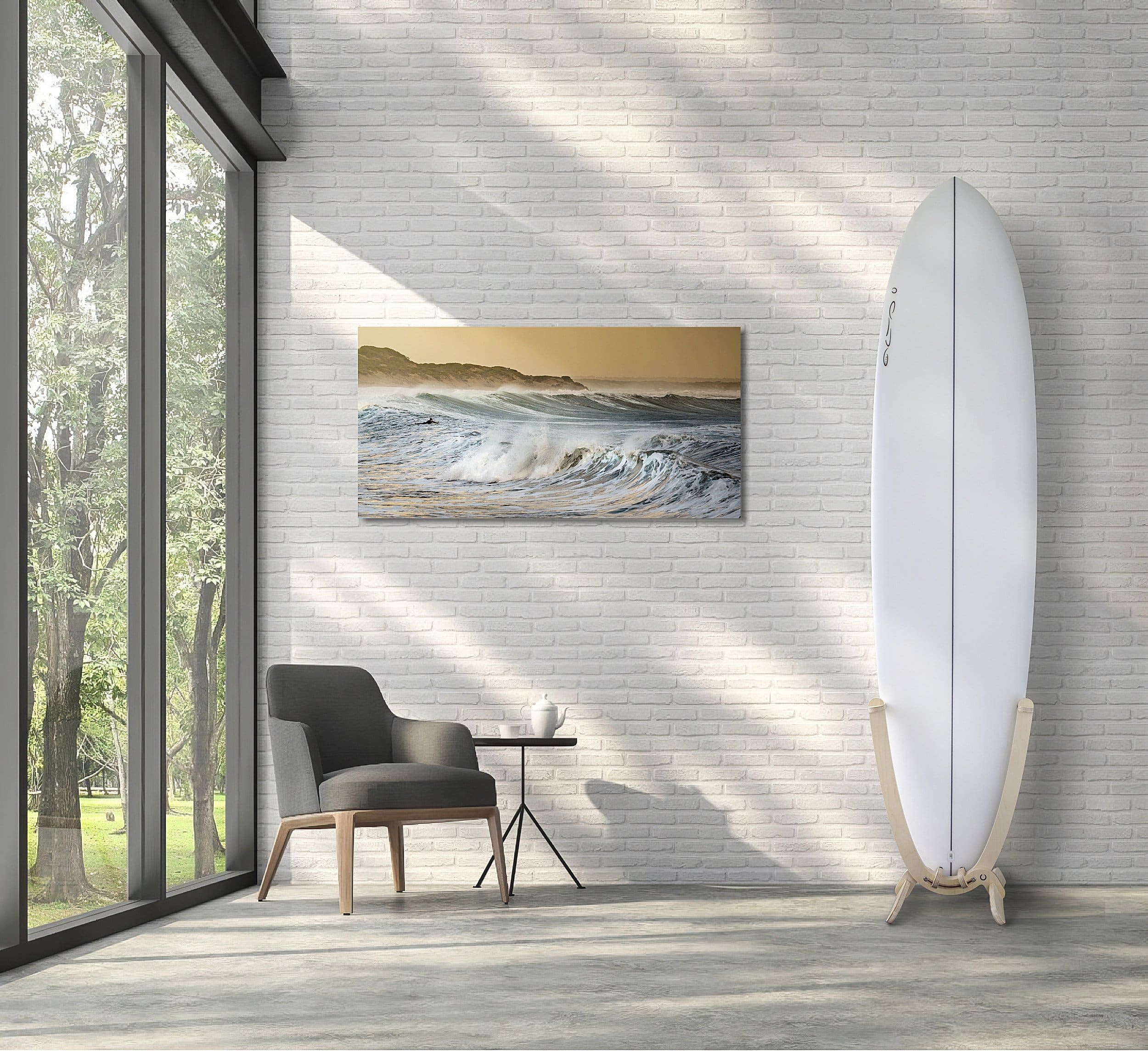 An all white Surfboard with a GLYD O decal on its nose, is mounted in a GLYD O surfboard rack. The rack with board is standing on a concrete floor to the right of the image against a white brick wall. There is a picture of waves on the wall to the left of the board and rack, a window on the left edge of the image and a chair and side table on the floor near the window. The room has much filtered light and shadows. The surfboard rack and surfboard really pop in this picture and the image gives you an idea of the feeling and vibe the GLYD O surfboard rack brings to a room. The Surfboard Rack is a Beautiful birchwood curved single surfboard rack in the shape of the tail of a surfboard, with hooks to hold the rails and tail and two slightly curved feet connected perpendicular to the upright curved arms. Image is taken from the front in relations to the vertical surfboard rack.