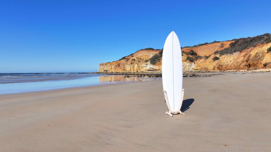 GLYD O Revolute Board Rack with surfboard mounted, standing on beach. Ocean and cliffs in background.