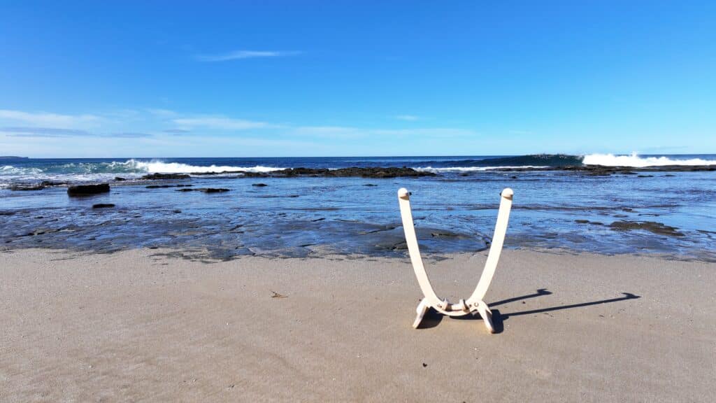 GLYD O Revolute Surfboard Display Rack, standing on beach. Reef, waves, ocean and sky in background.