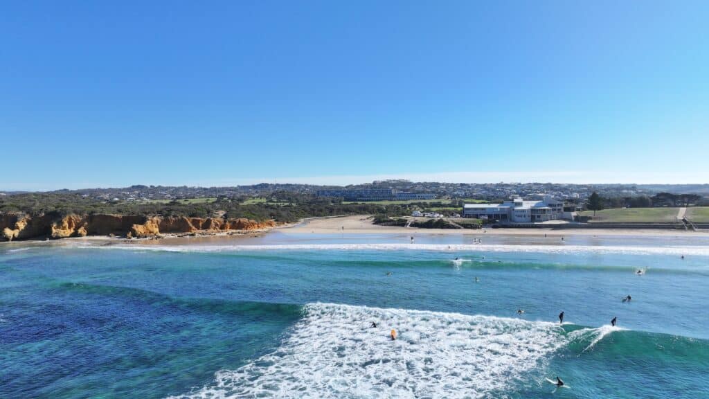 Aerial view of surfers riding clean waves at Torquay Beach, Victoria, with golden cliffs, clear blue water, and beachfront buildings under a bright blue sky.
