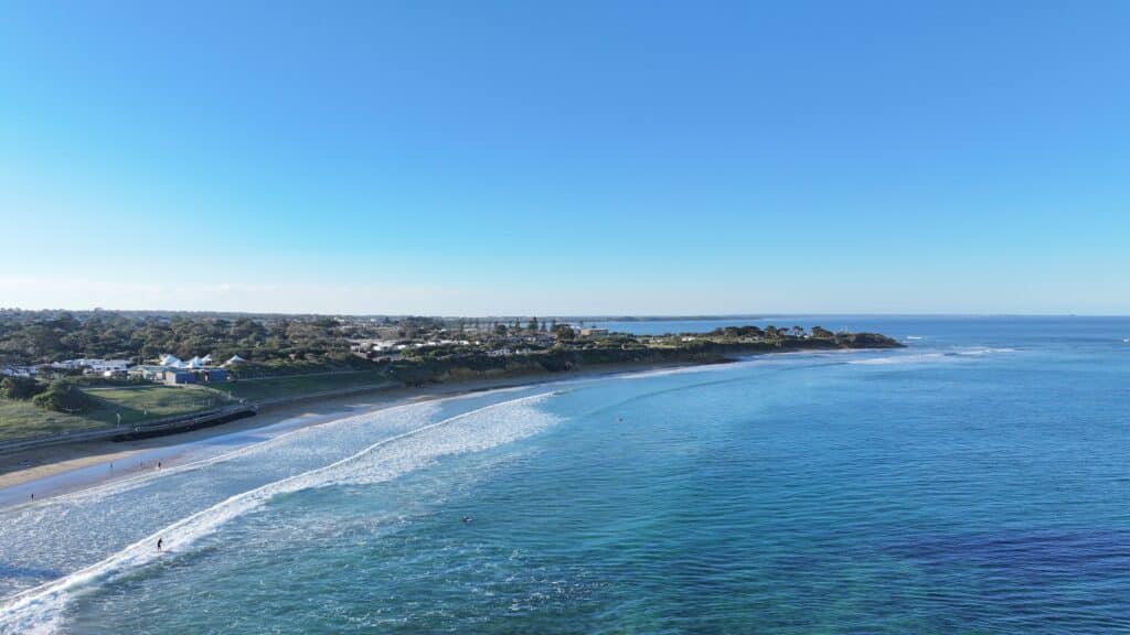 Aerial view of Torquay Beach, Victoria, stretching toward Point Danger with gentle waves, coastal vegetation, and clear blue skies over the ocean.