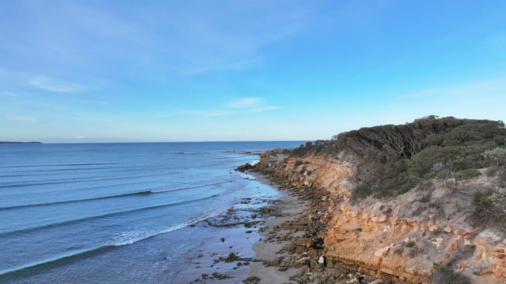 Point Roadnight eastern view showing ochre-colored rocky cliffs, small waves peeling right, coastal scrub vegetation, and Southern Ocean Bass Strait waters.