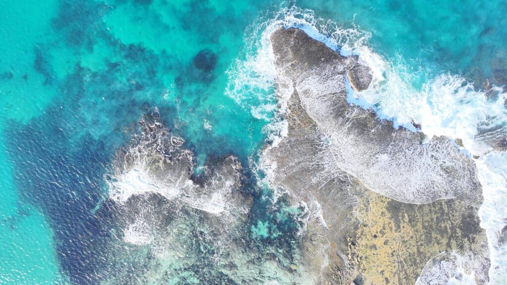 Aerial view directly above Point Roadnight reef showing turquoise waters surging over rocky formations creating white foam and revealing underwater reef structure.
