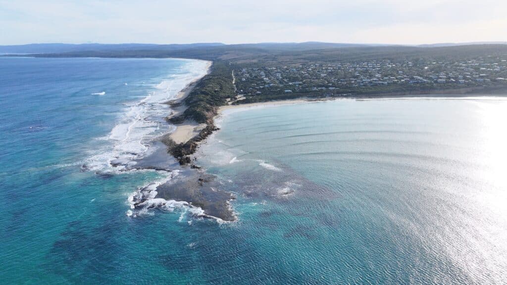 Aerial view of Point Roadnight from 120 meters showing rocky point, reef, waves, protected bay, open ocean, and coastal township from east facing west. Revolute Surfboard Display Stand on beach in distance.