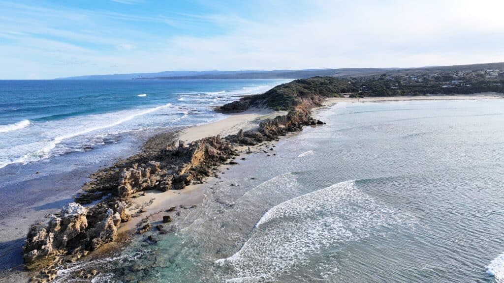 Aerial view of Point Roadnight from 50 meters showing detailed rocky point formations, waves breaking over reef, protected bay, and coastal township.