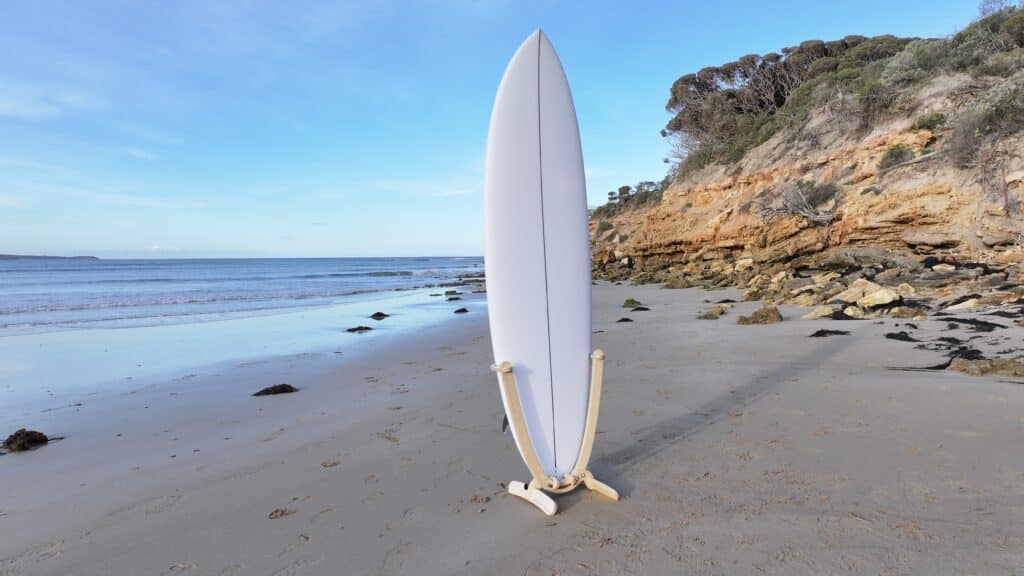 Revolute surfboard display stand holding white surfboard on sandy beach at Point Roadnight with ocean waves and coastal cliffs in background.