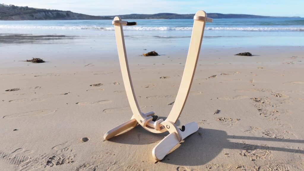 Close-up view of Revolute surfboard display stand on sandy beach at Point Roadnight showing wooden construction, stainless steel fasteners and foam inserts with ocean waves and coastline in background.