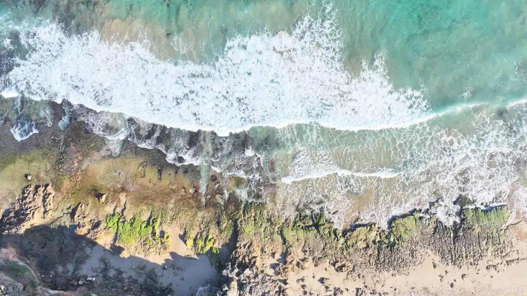 Aerial view from 120 meters above The Bluff beach showing reef formations, turquoise water, and white foam waves.