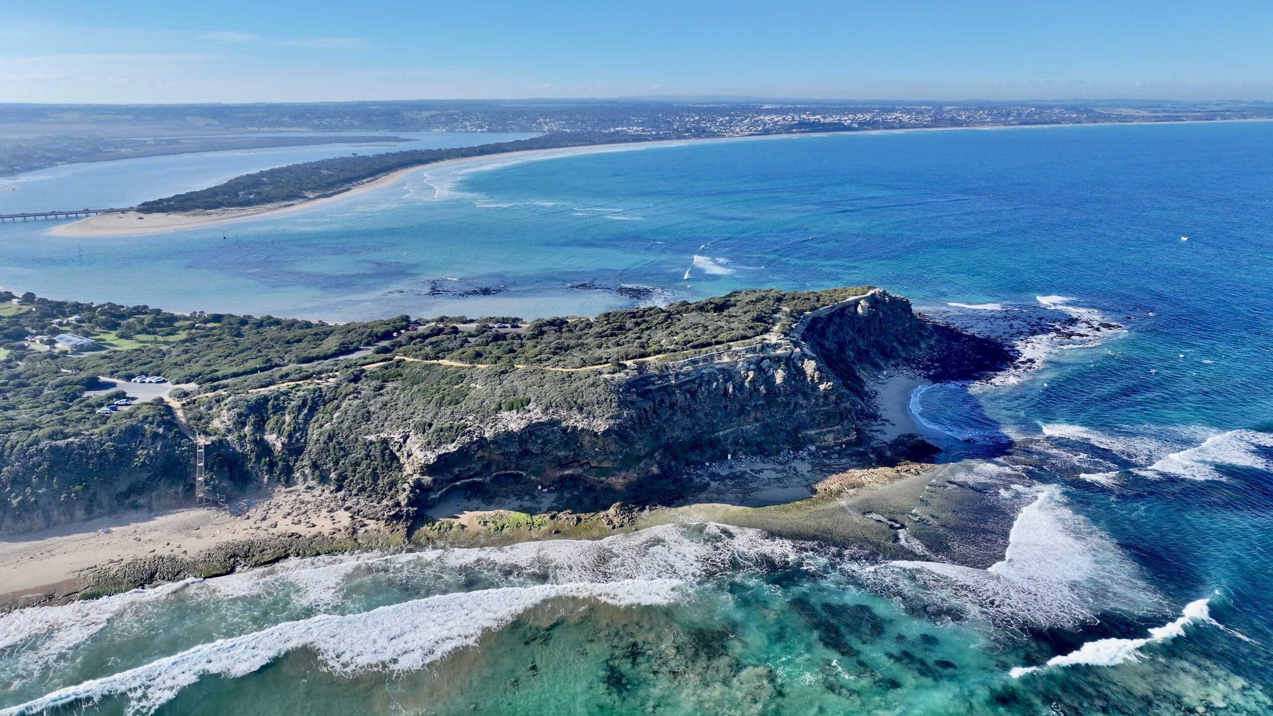 Aerial view of The Bluff Barwon Heads from above the surf break showing rocky cliffs, waves, and Ocean Grove coastline. Wooden board rack on beach.