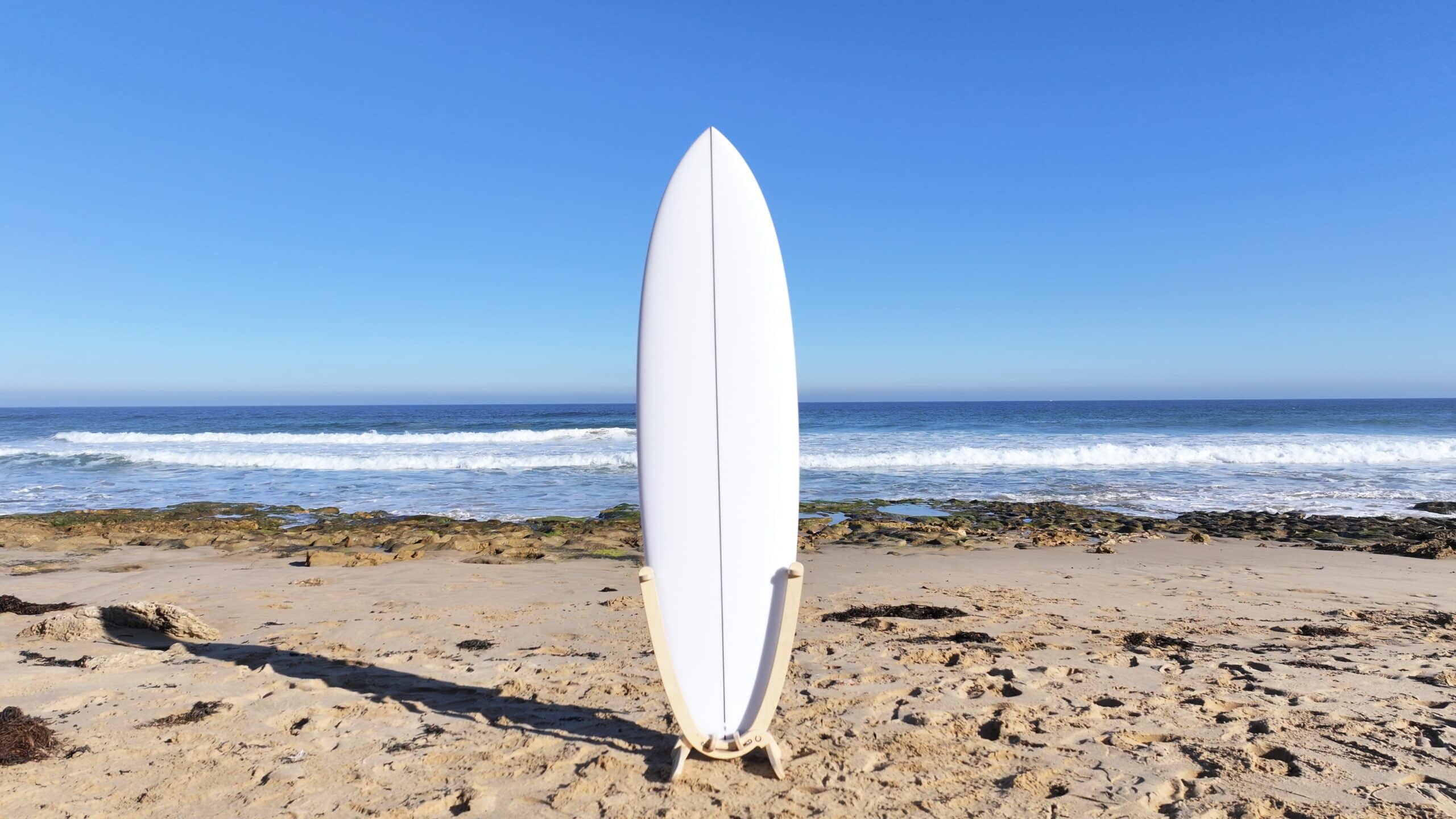 White surfboard displayed on wooden board rack at The Bluff Barwon Heads beach with ocean waves and blue sky.