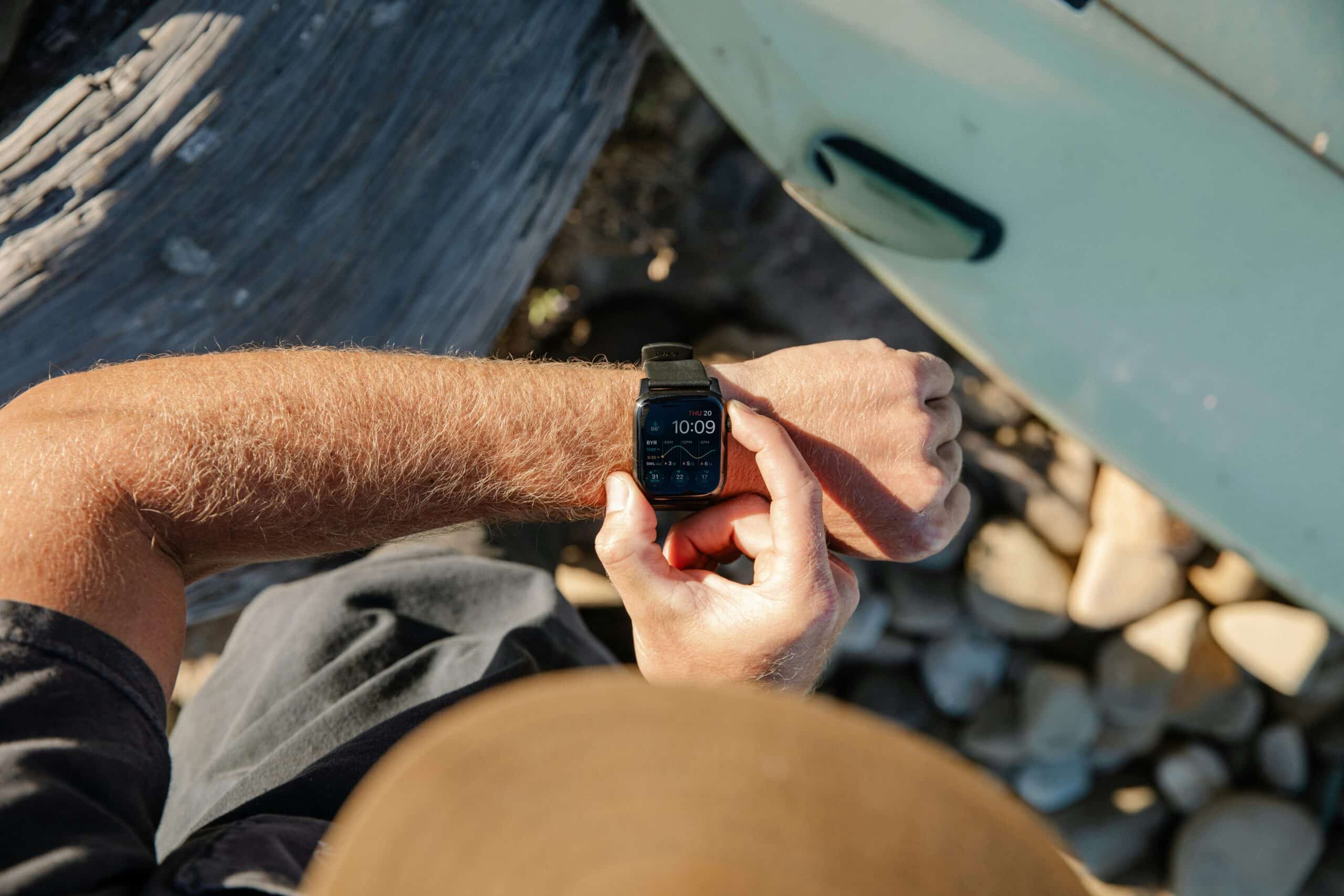 Person checking Apple Watch surf tracking app with surfboard in background on rocky shore